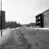 Österled med flerfamiljshus på båda sidor. Människor och bilar i rörelse. Vinter och snö.