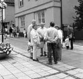 Det är höstmarknad på Stora torget. Några bekanta har träffats, intill Rådhusgården. Kvinnan till höger, i rutiga långbyxor, är Gudrun Sjökvist.
I bakgrunden ses Optikern. Glasögonbutiken ligger i hörnet Kapellgatan - Smedjegatan. Blomsterplantering på torget.