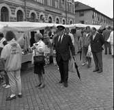 Höstmarknad. Människor i alla åldrar strosar bland torgstånden. Där erbjuds bland annat paraplyer.
En man i Frälsningsarméns uniform promenerar med en insamlingsbössa i ena handen. Paret bakom honom, klädda i jeansjackor, är Annika Sörén och Lars Sörén.
I bakgrunden ses 