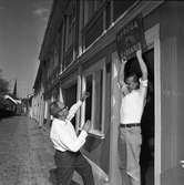 Bertil Levin och Yngvar Gunnarsson ställer iordning en diversehandel på Västerlånggatan. Nu ska de sätta upp en skylt med texten 