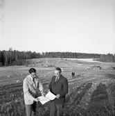 Kägleborgsbanan presenteras. 
Mannen till vänster kan vara Bengt Reander, Hedemora, Svenska Bilsportsförbundet.
Mannen till höger är Sture Gustafsson, sekreterare i Arboga SMK. (Sture blev senare ordförande, 1972).
Två män studerar en karta eller en ritning. Bakom dem ses den öppna yta som ska ställas iordning för motorsport, Kägleborgs motorstadion. Några bilar ses i bakgrunden.
Motocross
Mannen som är på väg till dem, är förmodligen Nils Lindgren, från Ålsäng (kallad Niklas i svängen).