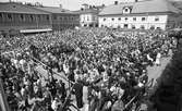 I Arboga firas riksdagens 550-årsjubileum. En stor publik har samlats på Stora torget. På bänkarna sitter riksdagsledamöterna.
I bakgrunden ses Lundborgska fastigheten med frisör och konditori Saga (observera publiken på taket!) och Duka-butiken.