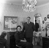 Evert Karlsson med hustrun Ingegärd. Fotot är taget på Trädgårdsgatan.
Mannen står och kvinnan sitter. I rummet ses en sekretär, en hög spegel, tavlor på väggarna.