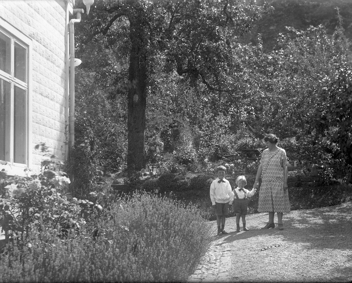 Calla Helena Sundbeck, A woman stands with children in her hand on the driveway to BergsEn 20. It is Anna-Greta Roman who with her children Sten and Lars. She holds the youngest, Lars, in her hand.