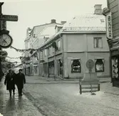 Västerås. Köpmangatan mot norr med korsningen Hantverkargatan.
Åkerboms ur hänger utanför butik, Guldbutik, Didners, blomsteraffär m.fl. 1963.