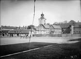 Nya torgets norra sida i Jönköping. Kristine kyrkas klocka visar nio på morgonen en vårdag år 1904. Torget anlades då den så kallade Breda hamnen, som var en del av kanalsystemet, fylldes igen på 1830-talet. Planket sattes upp efter en brand i augusti 1903.