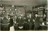 Interiör från en okänd handelsbod i Sundsvall. Butikspersonal och kunder. Vykort.
