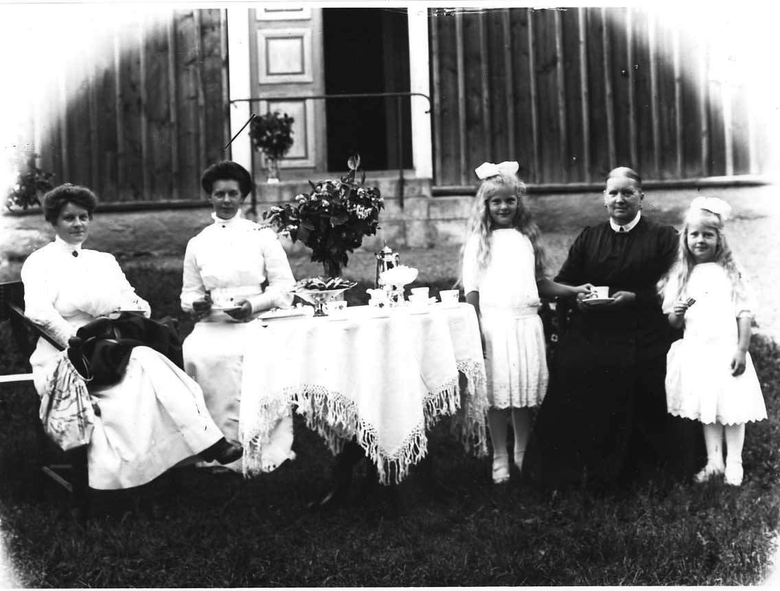 Calla Helena Sundbeck, A group picture with three women and two girls sitting and standing by a docked coffee table outdoors in front of Missionshus in Örserum, a red coloured building with lid list panel. On the left are two women in bright dresses, it is the siblings Agnes and Tyra Josefsson. On the right two girls in …