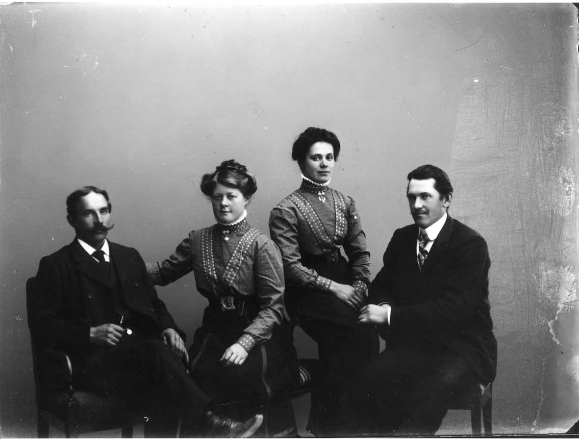 Calla Helena Sundbeck, Group image with two young women sitting between two men. The women are dressed in the same way with dark skirt and plaid blouse with spotted bands running from over shoulder oblique to the navel. The men wear a suit and a requirement.