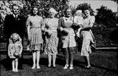 En samling människor står i solen i trädgården på Stora Ry med blommande buskar och lökväxter i en rabatt längs grusgången. Personerna är enligt bildförteckningen från vänster: Åke Johansson med dotter Eivor, som har en väska med en katt på, Signe, troligen någon som hälsar på från Amerika, Gerda och Linnea med dotter Solvig på armen.