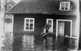 Översvämning.
Ett bostadshus står till viss del under vatten.
Två män utanför, varav den ene bär den andre på ryggen.