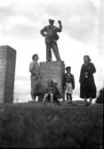 En grupp vuxna och barn, varav en man står på ett sten-fundament.