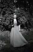 Signe Fredriksson (1901 - 1992) på väg till sin systersons bröllop cirka 1950. Hon är klädd i långklänning, kortärmad pälsjacka och har uppsatt hår. I bakgrunden finns buskar och växtlighet. Hon bodde i 