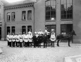 Framför brandstationen i kv. Kristina står ett antal uniformerade brandmän. Några sitter även på en redskapsvagn dragen av en häst. Vagnen köptes in 1913 och på kuskbocken sitter brandchef John Hj. Möller. I brandstationens ena fönster syns en person titta ut.