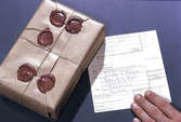 Seriebild H 9. En kund lämnar in ett litet paket med snören
förseglade med rött lack. På adresskortet anges att paketet, som
innehåller ett halsband, skall as-sureras för 100 kronor. Bilden är
tagen på poststationen Bromma 12.