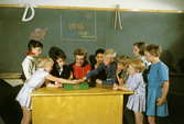 Seriebild L 4 a, alt 1. Skolsparsituation i klassrum i
Bergshamraskolan, Solna. Barnen lägger sina pengar i skolsparbössan
under lärarinnans överinseende. På svarta tavlan står uppmaningen.
