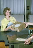 Seriebild H 16. Eva Sandström (fingerat namn) hämtar ett paket i
postluckan mot överlämnande av det kvitterade adresskortet, som
postkassören tar emot. Den arrangerade situationen är fotograferad i
paketkassan på postkontoret Farsta 1.