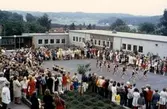 Examen och skolavslutning för elever på inre skolgården vid Brattåsskolan cirka 1970. Några elever i svarta gympadräkter gör en dansuppvisning. Vommedalsområdet (rött tegel) och Heljered ses i bakgrunden.
Relaterat motiv: A1501.
