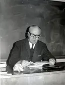 Folkskollärare och skolkantor Gunnar Alander sittandes (född i Göteborg 1900 - avliden 1979 i Kållered) vid en kateder i Kålleredskolan/Brattåsskolan cirka 1965. Bakom honom ses 