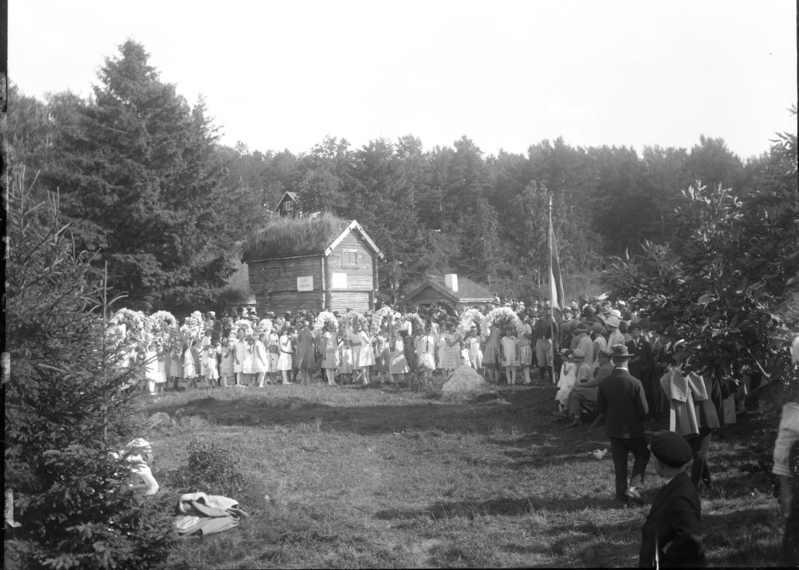 Calla Helena Sundbeck, Peoples gathering, children and adults at the Open-Air Museum at the Border Agency, it is Hembygdsfest. The children keep flower bows in their hands. They stand in front of the papermaker’s nest, whose loft shed has a sign: “Coffee tickets.”