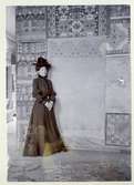 Stockholm. 
Kvinna stående framför vägg med bl.a. textilarbeten. 1901.