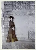 Stockholm. 
Kvinna stående framför vägg med bl.a. textilarbeten. 1901.