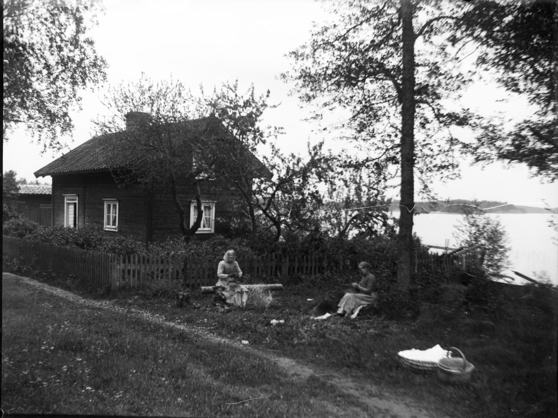 Calla Helena Sundbeck, Två kvinnor, den ena äldre med huckle, sitter intill en liten stuga vid en sjö. De ser ut att arbeta med något med händerna. Intill dem två korgare, den ena med en ljus duk.