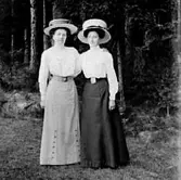 TVÅ KVINNOR I EN SKOGSGLÄNTA KLÄDDA I VIDBRÄTTADE HATTAR.