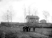 MINDRE GÅRD ELLER TORP MED FAMILJEN STÅENDE PÅ ÅKERN FRAMFÖR BOSTADSHUSET.