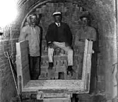 TRE MÄN SITTER PÅ TEGEL SOM STAPLATS I EN BRÄNNUGN. 
I FÖRGRUNDEN STÅR EN SPÅRBUNDEN VAGN FÖR TRANSPORT AV TEGLET IN I UGNEN.