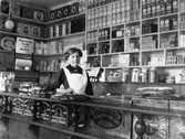 Bakom disken på Wessmans speceriaffär, Norra Strömgatan, står Olga Larsson klädd i vitt förkläde. Bakom henne hyllor fyllda med plåtburkar i olika storlekar och former.