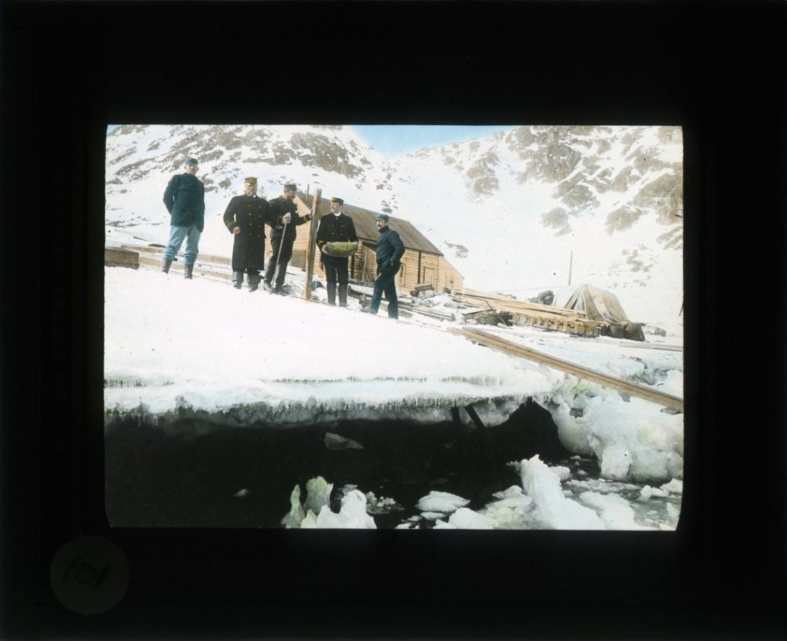 Carl Axel Wilhelm Stake, Five men stand on an ice edge below Pike’s house on Danish Island. They are from left: Andrée, Ehrensär, Norselius on skis, Celsing and Machuron. Coloured motif.