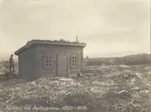 Första stugan vid Sylarna. Litet hus byggt av liggande timmer. Snedtak, två fönster och en dörr. En man i hatt står bakom stugan. Nyröjd mark runt huset, siluetter av fjäll i bakgrunden. 