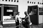 Två män i vardagligt samtal vid Esso i Veddige på 1950-talet. Till vänster står Karl-Erik Månsson och mannen med damcykeln heter Arne Tjärnmo. På pakethållaren har han en bunt tidningar. Bakom dem syns mackens skyltfönster och till höger portarna in till servicehallen.