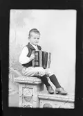 Ateljéporträtt - pojke klädd i folkdräkt sitter på en piedestal med ett dragspel i knät, Östhammar, Uppland