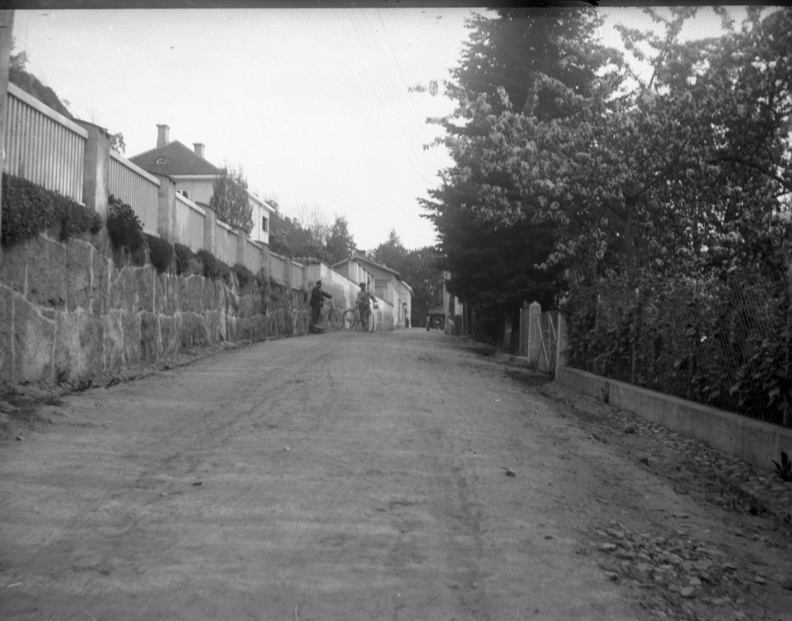 Unknown artist, Södra delen av Bergsgatan. Innanför hög stenmur och staket skymtar fastigheten Bergsgatan 8. På den ej belagda vägen står till vänster två män med keps vid cyklar. Längre bort till höger står en bil med nedfälld sufflet parkerad.