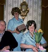 Avtackning av vårdbiträde Elsa Nilsson i Brattåshemmets matsal, maj 1979. Från vänster: Okänd man, föreståndare Aina Ekstedt samt Elsa Nilsson. På väggen i bakgrunden hänger en guldpendyl. Relaterade motiv: A1846 och A1855.