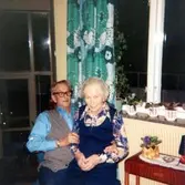 Sömmerskan Agnes Ekstedt (1898 - 1984) sitter i knäet på skräddaren Per Pettersson (1896 - 1981). De befinner sig i dagrummet på Brattåshemmet år 1979.