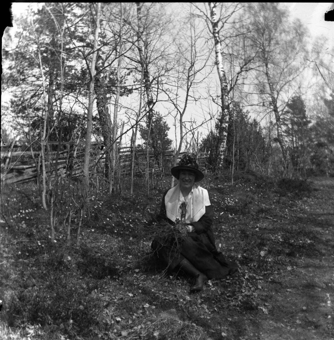 Unknown artist, An unknown woman sits in a hage in front of some diluted trees and a yard. She possibly holds a bundle of rice in her hands. She wears her hat coh on a handrail, probably in the steps of the Border Guard, in evening light over Vättern. She wears a hat and possibly a dark coat with a large light col…