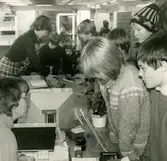 Biblioteksassistent Ingrid Hansson (ståendes till vänster) vid utlåningsdisken på Kållereds bibliotek 1980-tal.