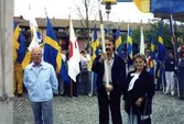 Valmöte eller 1 maj-möte på Alberts torg i Mölndal, 1980-tal. Från vänster: Rolf Andersson, ordförande i Kållereds S-förening, Okänd samt Johanna Karlsson, Kållered. Flertalet människor står bakom dem och håller i Svenska flaggor. Längre bort i bakgrunden skymtas lägenheter utmed Bergmansgatan.