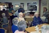 Sångstund i Brattåsgårdens hobbylokal med Stig-Ove Dahlgren på gitarr och Ivar Andersson på fiol, cirka 1990. Längst till höger sitter Stina Svensson (i vit kjol) och snett framför henne vid bordet sitter Rut Göransson (i mörk klänning). Övriga är okända.