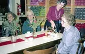 Försäljning av hantverk i pensionärernas hobbylokal på Våmmedalsvägen år 1982. Från vänster: Valborg Smitt 1901 - 1986), Hulda Olsson, Helga Svensson (1910 - 1985) samt okänd kvinna. På hyllorna i bakgrunden ligger olikfärgade mattgarner staplade för att användas till vävning.