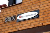Skylt med husnummer och skylt med MölndalsBostäders logotyp på husfasaden. Byggnadsdetalj från huset med adress Bergmansgatan 8-10 i Mölndals Centrum. Fotografi taget den 26 februari 2018. Byggnadsdokumentation inför rivning.