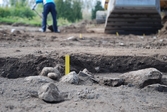 En arkeolog arbetar vid grävmaskinen i bakgrunden och i förgrunden syns en anläggning med stenar markerad med en gul pinne. Foto taget vid en arkeologisk undersökning i Gränna 2009.