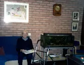 En okänd man sitter i en soffa och bläddrar i en bok, Brattåsgården (Streteredsvägen 5) cirka 1990. Vid sidan av mannnen står hans rullator samt ett akvarium. Bakom mannen är en gul tegelvägg.