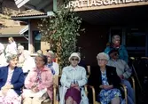 Midsommar utanför Brattåsgården, cirka 1990. Främre raden från vänster: 1. Gunvor Johansson, 2. Okänd dam (rosa jacka), 3. Ingrid Rosén (vit hatt), 4. Helga Posse (blommig klänning), 5. Okänd man i solglasögon, 6. Okänd dam i grön jacka. I bakgrunden skymtar Eva, Oskar och Svea Jonasson samt Elsa Olausson.