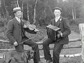 Edard Johansson med fiol, och brodern Klas Johansson med dragspel. Männen sitter på ett stängsel vid en hage med skog i bakgrunden. En hund står bredvid dem.