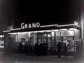 Biobesökare eller flanörer utanför entrén till biograf Grand,  kvarteret Prosten, Varberg. Neonskylten 
