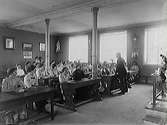 Interiör från A-salen på Katrinebergs folkhögskola. De kvinnliga eleverna sitter vid skolbänkar och rektor Ludvig de Vylder står lutad vid en av de främre. Det är troligen en klass från lanthushållsskolan. 1910-talet.